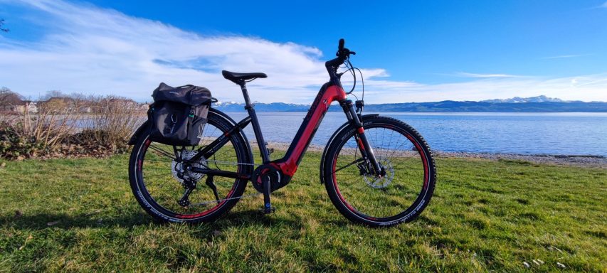 Rotes Fahrrad mit Gepäckträger auf grünem Gras neben einem Gewässer und Bergen im Hintergrund.